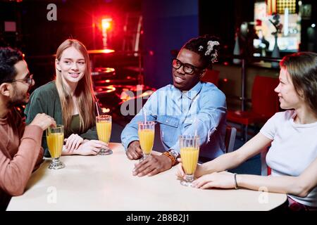 Two young intercultural couples in casualwear having orange juice while sitting by table in cafe after playing bowling and chatting Stock Photo