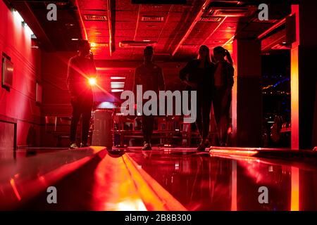 Silhouettes of four young people standing by bowling alley in dark room while one of guys going to throw ball Stock Photo