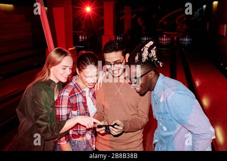 Group of happy young intercultural friends scrolling through their photos in smartphone after playing bowling in leisure center Stock Photo