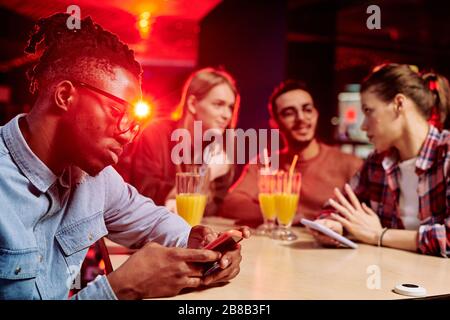 African guy scrolling in smartphone while sitting by desk in cafe on background of friends having drinks and chatting Stock Photo