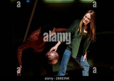 Young man looking at his cheerful pretty girlfriend with bowling ball while teaching her how to play it by alley at leisure center Stock Photo