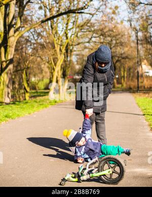 POZNAN, POLAND - Mar 15, 2020: Woman pulling up a small boy that fell on a footpath with a running bike in the Jan Pawla park on a sunny day in the wi Stock Photo