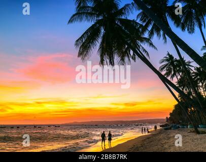 Sunset on the beach with silhouette couple, tilted coconut trees, long sandy beaches and beautiful golden sky and romantic for the weekend resort. Stock Photo