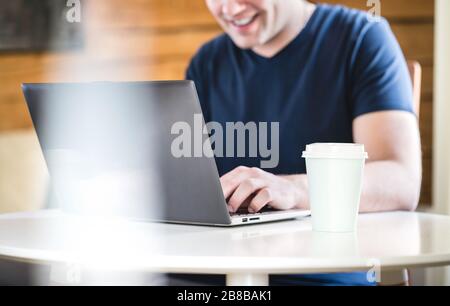 Happy man using laptop with take away coffee cup on table. Smiling person with computer home or in cafe. Stock Photo