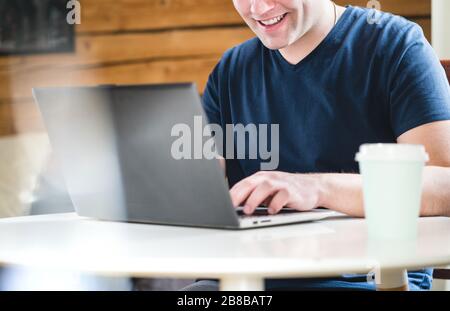 Happy man using laptop home or cafe. Smiling person with computer and take away coffee cup on table. Stock Photo