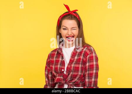 Portrait of optimistic pinup girl in checkered shirt and headband winking to camera with crazy naughty expression, showing tongue out, making faces bl Stock Photo
