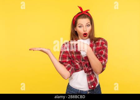 Crazy product advertising. Portrait of amazed pretty pinup girl in checkered shirt and headband pointing at copy space on her palm, empty place for co Stock Photo