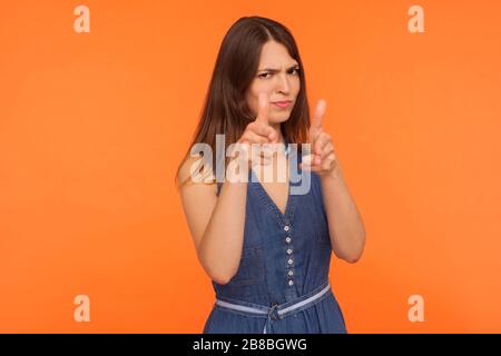 Gun gesture. Annoyed brunette woman in denim dress pointing finger pistol to camera, threatening to shoot, looking displeased angry and dangerous. ind Stock Photo