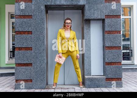 The full-size photo of a business lady in a colorful pantsuit with a pink bag. The model is posing for a photographer near the elevator entrance. Stock Photo