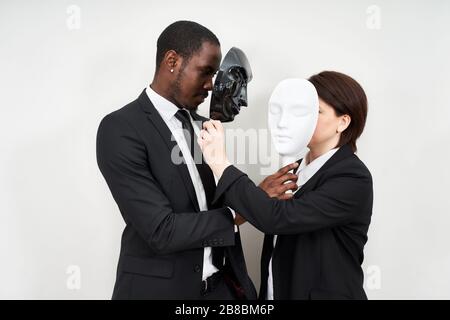 African man and Caucasian woman wearing black suits hiding face with plastic masks Stock Photo