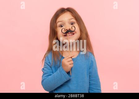 Portrait of cheerful little girl holding paper mustache and smiling to camera, carefree child playing, making funny face, wearing masquerade accessory Stock Photo