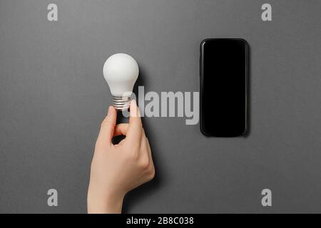 Creative top view flat lay of female hand holding light bulb and modern smartphone mockup composition with copy space on grey background minimal style Stock Photo