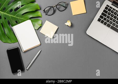 Workspace with modern smartphone, laptop, pen, monstera leaves, eyeglasses and notebook on grey background minimal style. Flat lay, top view office ta Stock Photo