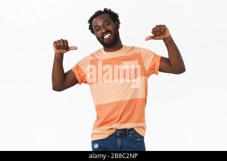 Confident, cheeky young musculine bearded african-american guy in striped t-shirt looking manly proudly pointing himself with raised thumbs, tilt head Stock Photo