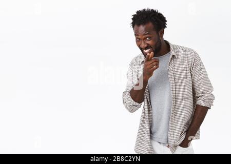 Cheerful, funny and cheeky young african-american bearded man in casual clothes, pointing finger camera, gotcha gesture and smiling sly, hinting Stock Photo