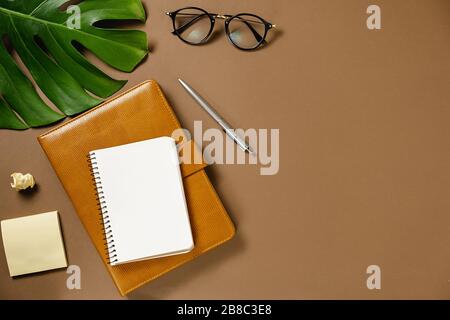 Workspace with pen, green monstera leaves, eyeglasses and two notebook on brown background minimal style. Flat lay, top view office table desk. Templa Stock Photo