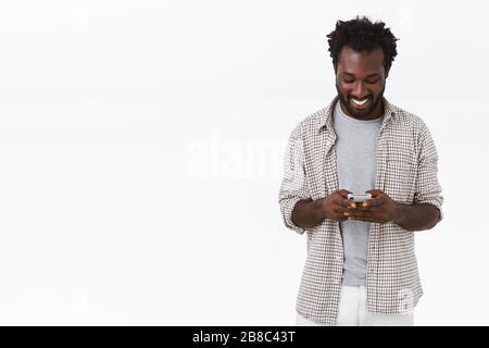 Modern young happy african-american bearded man in casual plain shirt, holding mobile phone, messaging via smartphone or use app to watch funny videos Stock Photo