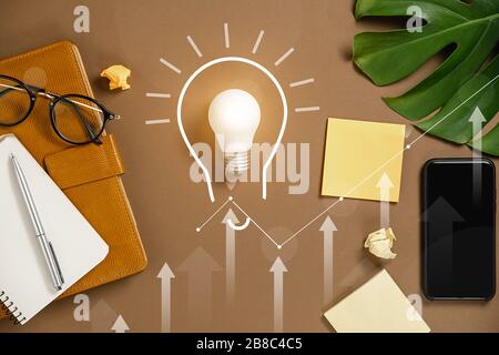 Ideas inspiration concepts with lightbulb, modern smartphone, pen, monstera leaves, eyeglasses and notebook with copy space on brown background minima Stock Photo