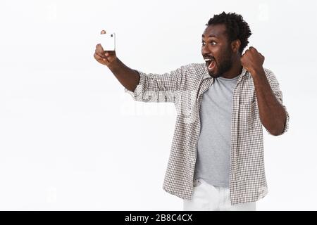 Guy watching online stream with smartphone application, triumphing, cheerfully celebrating team won, watching amusing game mobile phone, fist pump and Stock Photo