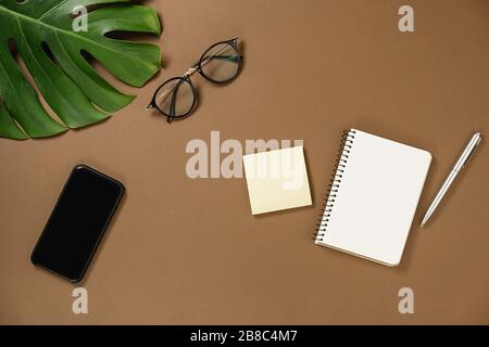 Creative flat lay, top view office table desk. Workspace with pen, monstera leaves, eyeglasses, notebook, smartphone with copy space on brown backgrou Stock Photo