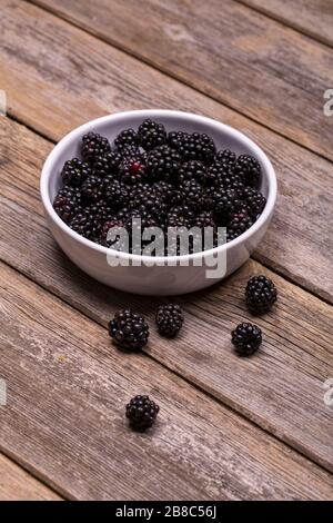 Freshly picked blackberries in a white ceramic bowl, on an old, wooden table. Some of the blackberries have spilled out onto the wood surface. Stock Photo