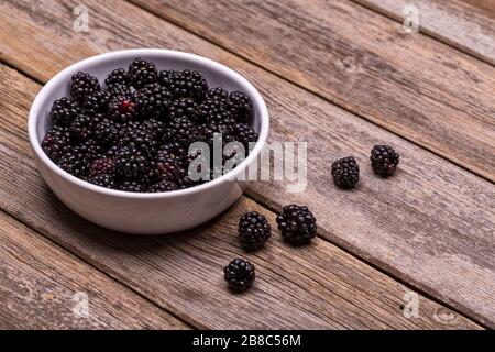 Freshly picked blackberries in a white ceramic bowl, on an old, wooden table. Some of the blackberries have spilled out onto the wood surface. Stock Photo