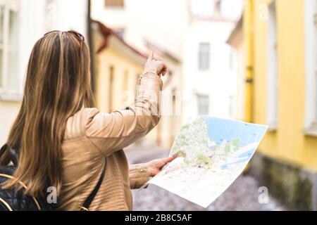 Tourist with map in the city. Woman pointing at right direction with finger. Back view of happy traveler using tourism guide. Person with backpack. Stock Photo