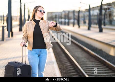 Woman looking at the time and watch while waiting for train. Smiling and happy female traveler standing at station and platform. Stock Photo
