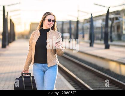 Woman walking in train station and using smartphone. Passenger buying electronic digital ticket with mobile phone. Happy lady pulling suitcase- Stock Photo