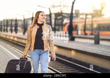 Lady pulling suitcase and luggage in train station. Woman walking in platform and holding ticket and passport. Happy female traveler. Stock Photo