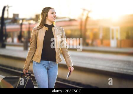 Happy woman pulling suitcase in train station. Smiling lady with baggage and luggage arrived to destination. Tourist on vacation. Stock Photo