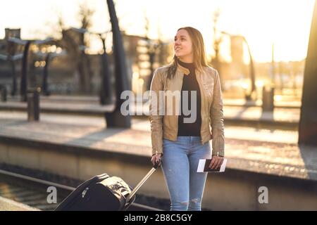 Female traveler standing in train platform at sunset. Smiling woman waiting at station with suitcase, baggage and luggage. Travel lifestyle. Stock Photo