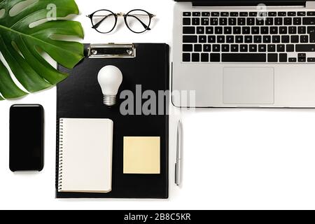 Modern workspace with light bulb, laptop, pen, monstera leaves, eyeglasses, clipboard, smartphone and notebook with copy space on white background min Stock Photo