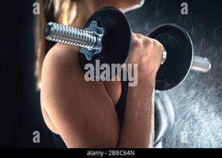 Fitness athlete doing bicep curls with dumbbell. Fit woman working out in gym. Sweat or water splashing. Hard workout, determination, effort. Stock Photo