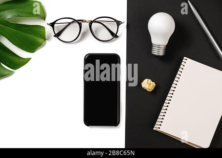 Financial planning table top with monstera leaves, pen, notebook, modern smartphone, eyeglasses and light bulb on white and black background minimal s Stock Photo