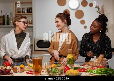 Group of young beautiful women standing in the kitchen cutting vegetables for salad and talking during cooking Stock Photo