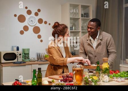 Young multiethnic couple standing at the table and cooking meal together in the kitchen they preparing for the dinner with friends Stock Photo