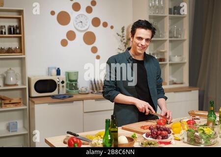 Portrait of young man standing at kitchen table and smiling at camera while cutting tomatoes on cutting board in the kitchen Stock Photo