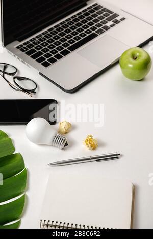 Minimal home office desk. Side view flat lay of modern workspace with light bulb, laptop, pen, monstera leaves, eyeglasses, fresh apple, smartphone an Stock Photo