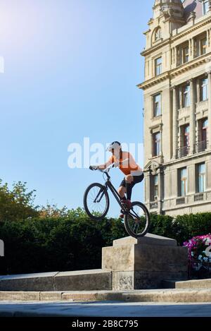 Side view of a fearless man showing trick on bicycle in city center. Young cyclist in helmet and orange shirt riding on architecture background. Concept of extreme. Stock Photo