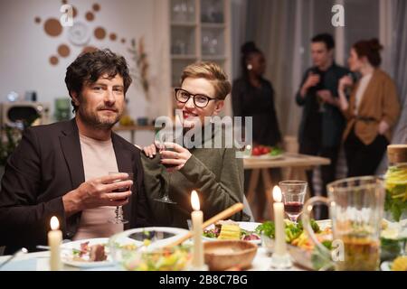 Portrait of young couple smiling at camera while sitting at the table and drinking wine during the party Stock Photo