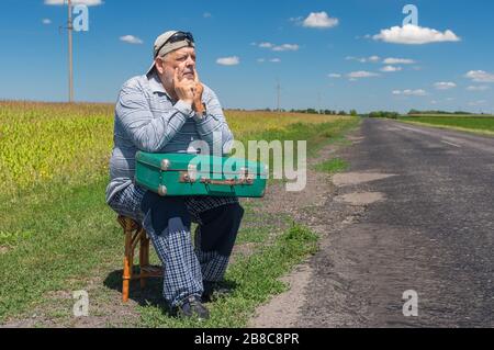 Portrait of Caucasian senior having short rest on a roadside sitting on a whicker stool with an ancient green suitcase Stock Photo