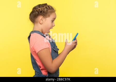 Side view of shocked little girl reading surprising message on cellphone, chatting on social network, amazed by good mobile application for children. Stock Photo