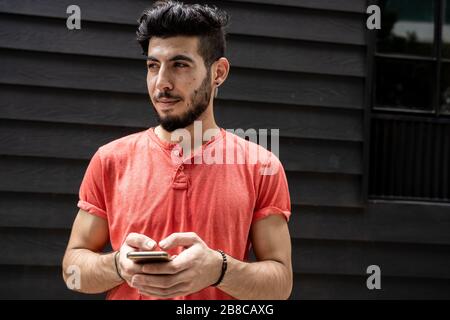 Young Arabian man dressed in pink tshirt using a mobile phone on the street in front of a blue wooden building Stock Photo