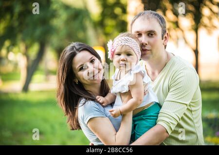 Happy young parents walking, playing and kissing their small daughter in park on summer clear day. Happy family time together concept Stock Photo