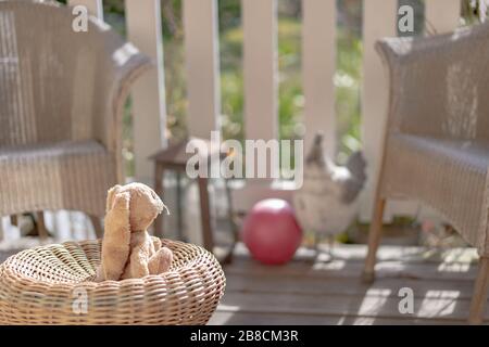 Stuffed toy easter bunny sitting on a straw chair on the veranda, looking to the sun. High angle view, close up, concept. Stock Photo