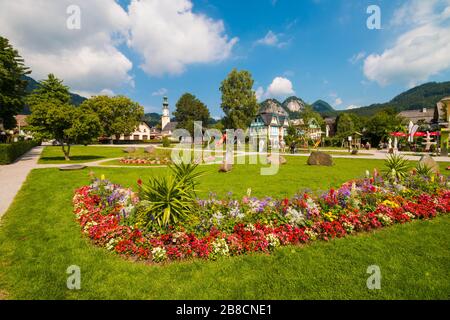 St.Gilgen, Austria - July 16, 2019: Park with green lawn and beautiful flowerbed in austrian alpine town, St. Giles church and mountains on the backgr Stock Photo