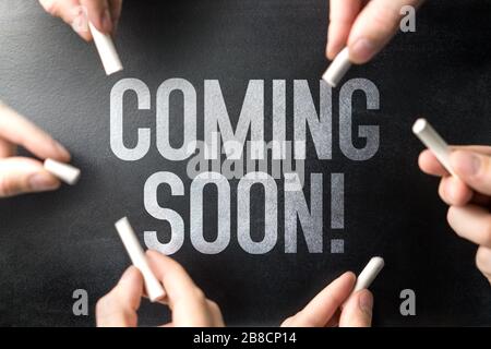 Coming soon text written on chalkboard sign and blackboard banner by many hands. Website or web page under construction or new announcement. Stock Photo
