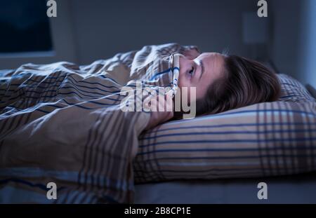 Scared woman hiding under blanket. Afraid of the dark. Unable to sleep after nightmare or bad dream. Awake in the middle of the night in bedroom. Stock Photo