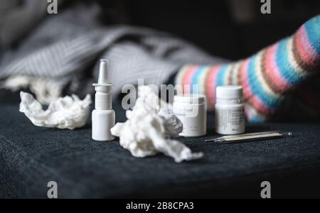 Flu and cold medicine. Sick woman with nasal spray, warm woolen socks, tissues and thermometer resting on couch at home. Ill person lying on sofa. Stock Photo